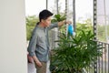 Attractive Young Man on Apartment Balcony Watering Plants in Box from Blue Watering Can on Sunny Day