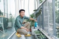 Attractive Young Man on Apartment Balcony Watering Plants in Box from Blue Watering Can