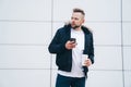 Attractive young man in casual clothers with coffee and phone in his hands standing on office building background. Royalty Free Stock Photo