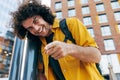 Attractive young man with curly hair, smiling broadly, looking, and pointing to the camera in the city street. Happy student male Royalty Free Stock Photo