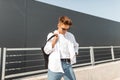 Attractive young man model in summer white fashionable clothes in sunglasses with a cloth black bag stands on a sunny day Royalty Free Stock Photo