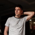 Attractive young man in a stylish striped t-shirt straightens hair standing in a dark room. Handsome guy model posing in the room.