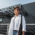 Attractive young man in stylish white shirt in fashionable sunglasses in trendy jeans with a cloth black bag posing on a sunny day Royalty Free Stock Photo