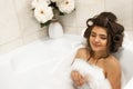 Attractive young relaxed brunette woman in hair curlers enjoying her bath with foam in bright bathroom