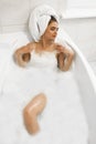 Attractive young relaxed brunette woman wearing white towel on her head enjoying her bath with bubbles in bright bathroom