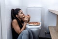 Attractive young and sad bulimic young woman feeling guilty and sick eating while sitting on the floor next to the toilet in