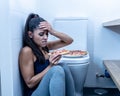 Attractive young and sad bulimic young woman feeling guilty and sick eating while sitting on the floor next to the toilet in Royalty Free Stock Photo