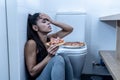 Attractive young and sad bulimic young woman feeling guilty and sick eating while sitting on the floor next to the toilet in Royalty Free Stock Photo