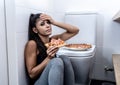 Attractive young and sad bulimic young woman feeling guilty and sick eating while sitting on the floor next to the toilet in