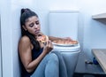 Attractive young and sad bulimic young woman feeling guilty and sick eating while sitting on the floor next to the toilet in Royalty Free Stock Photo