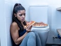 Attractive young and sad bulimic young woman feeling guilty and sick eating while sitting on the floor next to the toilet in Royalty Free Stock Photo