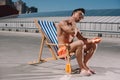 attractive young shirtless man sitting on sun lounger and applying sunscreen lotion Royalty Free Stock Photo