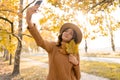 Attractive young smiling woman walking in autumn park taking selfie with leaf using smartphone, wearing sweater and hat Royalty Free Stock Photo