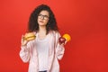 Attractive young student woman makes a choice between healthy and unhealthy foods, apple and hamburger isolated on red background