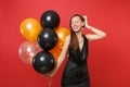 Attractive young woman in black dress celebrating, putting hand on head holding air balloons isolated on red background Royalty Free Stock Photo