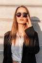 Attractive young woman in black stylish blazer straightens sunglasses and enjoys bright sunshine. Beautiful pretty urban girl in Royalty Free Stock Photo