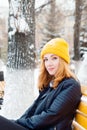 Attractive young woman with blue eyes and blond hair in a yellow knitting hat sitting on a yellow bench in the winter park Royalty Free Stock Photo