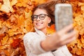 Attractive young woman with a cute smile photographs themselves on the phone with an orange maple leaf. Happy hipster girl in a Royalty Free Stock Photo