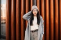 Attractive young woman in fashionable coat in stylish knitted hat in trendy glasses in sweater poses near vintage metal wall in Royalty Free Stock Photo