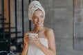 Attractive young woman puts moisturizing cream on face after showering, smiles broadly, wrapped in towels, drinks coffee, poses Royalty Free Stock Photo