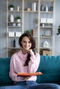 Attractive young woman is reading a book, looking at camera and smiling sitting on a cozy sofa Royalty Free Stock Photo