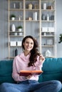 Attractive young woman is reading a book, looking at camera and smiling sitting on a cozy sofa Royalty Free Stock Photo