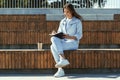 Attractive young woman sits on public park bench, writes in black notebook. Brunette girl dressed in denim clothes Royalty Free Stock Photo