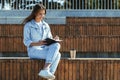 Attractive young woman sits on public park bench, writes in black notebook. Brunette girl dressed in denim clothes Royalty Free Stock Photo