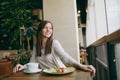 Attractiveyoung woman sitting alone near big window in coffee shop at table with cup of cappuccino, cake, relaxing in