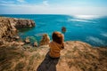 Attractive young woman stands on a high cliff overlooking the rocky cliffs arches on the beach and turquoise sea water on the coas Royalty Free Stock Photo