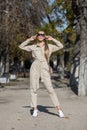 Attractive young woman in sunglasses and overalls similar to the uniform posing in autumn park with hands raised Royalty Free Stock Photo