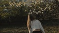 Attractive young woman in sweater, jeans is dance and spinning among blossom apple tree. Sunset, spring orchard and