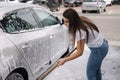 Attractive young woman washing her car with shampoo and brushes. Female washes automobile with foam and water outside on Royalty Free Stock Photo