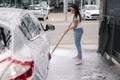 Attractive young woman washing her car with shampoo and brushes. Female washes automobile with foam and water outside on Royalty Free Stock Photo