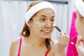 Attractive young woman wearing pink top and white headband, applying oat mixture to face using brush, looking in mirror Royalty Free Stock Photo