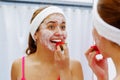 Attractive young woman wearing pink top and white headband with cream on face, eating strawberry, looking in mirror Royalty Free Stock Photo