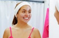 Attractive young woman wearing pink top and white headband, half of face covered with cream, looking in mirror smiling Royalty Free Stock Photo