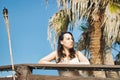 An attractive young woman in a white bikini leaning on a balcony railing in front of a palm tree looking into the distance Royalty Free Stock Photo