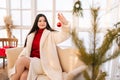 Attractive young woman in white fur coat holding Christmas red ball sitting on sofa in light cold summer house room Royalty Free Stock Photo