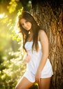 Attractive young woman in white short dress posing near a tree in a sunny summer day. Beautiful girl enjoying the nature