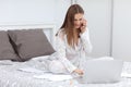 Attractive Young Woman Working from Home - Female Entrepreneur Sitting on Bed with Laptop Computer, Paperwork and tolking on the Royalty Free Stock Photo