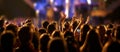 Audience with hands raised at a music festival and lights streaming down from above the stage.