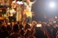 Audience with hands raised at a music festival and lights streaming down from above the stage.