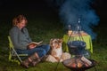 Australian Shepherd dog is lying by the campfire, the kettle with food is cooking and steaming, The woman is reading a Royalty Free Stock Photo