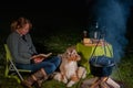 Australian Shepherd dog is lying by the campfire, the kettle with food is cooking and steaming, The woman is reading a Royalty Free Stock Photo