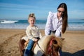 Authentic family enjoying happy time on the beach. Loving caring mother teaching her lovely child to rise a horse pony