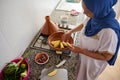 Authentic Muslim woman in hijab, standing by stove on kitchen counter and stacking potato slices in a tagine clay pot