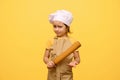 Studio portrait of amazed little girl, chef pastry, in white cap and beige apron, hoding rolling pin, isolated on yellow Royalty Free Stock Photo
