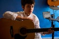 Handsome teenage boy playing guitar in the modern music studio, looking confidently at camera, sitting against a blue Royalty Free Stock Photo