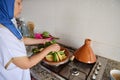 Authentic Muslim woman with head covered in hijab, putting fresh green beans on vegetables, steaming in tagine clay pot